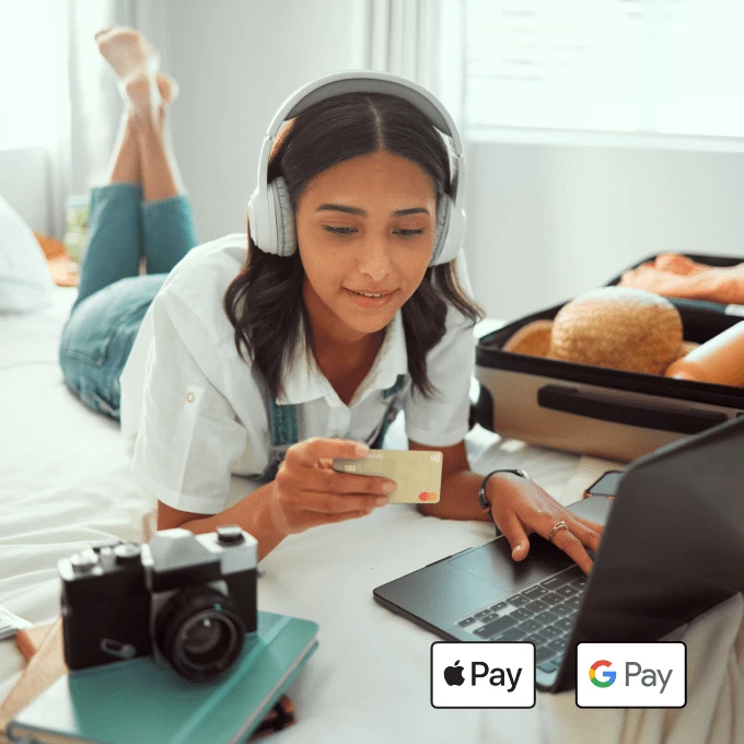 Mädchen liegt auf Bett mit Kopfhörern auf. Vor ihr liegt ein Laptop und sie hat die TF Mastercard Gold in der Hand. Auf dem Bett liegt außerdem bereit ein Koffer und ein Fernglas.